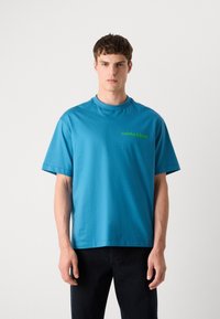 Μπλε βαμβακερή μπλούζα με στρογγυλή λαιμόκοψη και κοντά μανίκια, με πράσινο κείμενο "calvin klein" στο στήθος. Απλός σχεδιασμός, κανονική εφαρμογή.