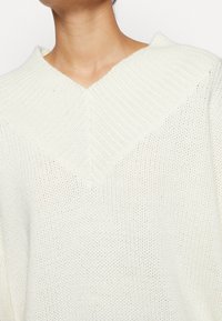 Cremfarbener, gestrickter Pullover mit V-Ausschnitt, der ein strukturiertes Rippenmuster und einen lockeren Schnitt aufweist und aus weichem, atmungsaktivem Stoff gefertigt ist.