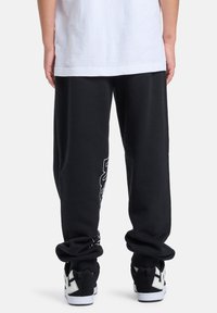 Schwarze Jogginghose mit lockerem Sitz und weißen Grafiken am linken Bein. Kombiniert mit schwarz-weißen Turnschuhen.