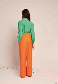 Zelena bluzica z dolgimi rokavi z napihnjenimi rokavi v kombinaciji z oranžnimi širokimi hlačami visokega pasu, ki imajo zavihek na robu.