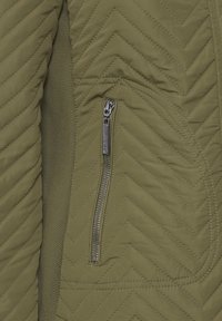Tissu matelassé vert olive avec des surpiqûres en zigzag et une poche verticale zippée argentée avec un tire-zip sur le côté droit.
