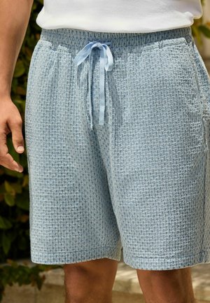 Hellblaue, strukturierte Strickshorts mit Vordertaschen und gebundener Kordel im Taillenbereich, getragen von einer Person in weißem Oberteil.