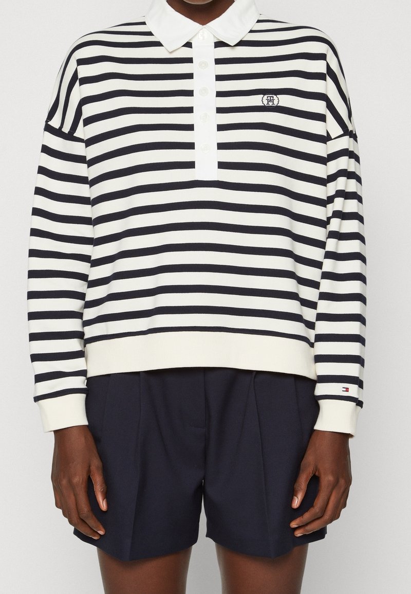 Sweterek polo w paski w kolorze kremowym i granatowym, z białym kołnierzykiem i guzikami, haftowanym logo na piersi oraz ściągaczami przy mankietach.