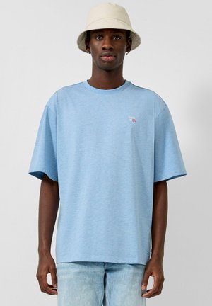Homme portant un t-shirt oversize bleu clair avec un petit logo, un jean clair en denim et un chapeau beige type bob, debout devant un fond gris uni.