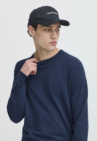 Giovane uomo in un maglione blu navy e un berretto nero con la scritta "Casual Apparel", che guarda di lato mentre sistema il colletto del suo maglione.