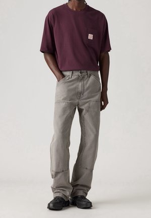 Homme portant un jean gris ample avec des détails de couture sur les poches, un t-shirt violet foncé avec une poche poitrine et des baskets noires, debout les mains dans les poches.