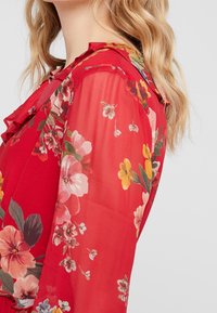Blouse florale rouge en tissu transparent, avec un col froncé et divers motifs de fleurs colorées. Matière légère au silhouette fluide.