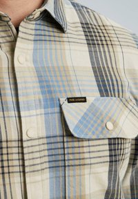 Chemise à carreaux beige, bleu et noir. Manches longues, col boutonné et poche poitrine avec une étiquette PME Legend.