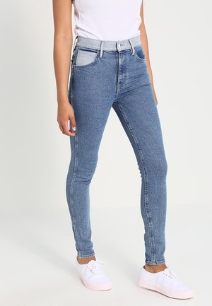 Mujer con jeans ajustados azules con cinturilla gris y zapatillas blancas, de pie frente a un fondo liso.