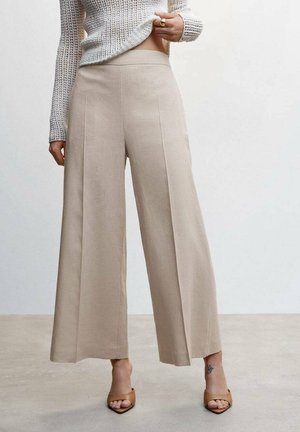 Kobieta nosząca beżowe szerokie spodnie z wysokim stanem, krótki sweter z dzianiny, beżowe sandały na obcasie typu open-toe oraz mały tatuaż na kostce lewej nogi.