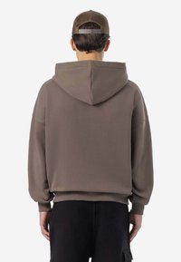Brauner übergroßer Hoodie mit Kordelzugkapuze und gerippten Bündchen, kombiniert mit schwarzen Cargohosen. Der Stoff wirkt glatt und leicht.