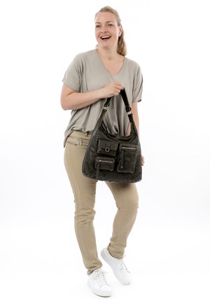 Femme debout, souriante, tenant un sac à main noir avec plusieurs poches avant, portant un pantalon beige, un haut gris et des baskets blanches.