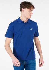 Polo blu realizzato in tessuto strutturato, con colletto classico, maniche corte e un logo circolare sul lato sinistro del petto.