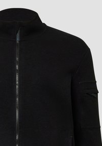 Zwarte ritsjack met hoge kraag, gemaakt van zachte stof; beschikt over ribbelmanchetten, een ritsfront en een klein zakje op de mouw.