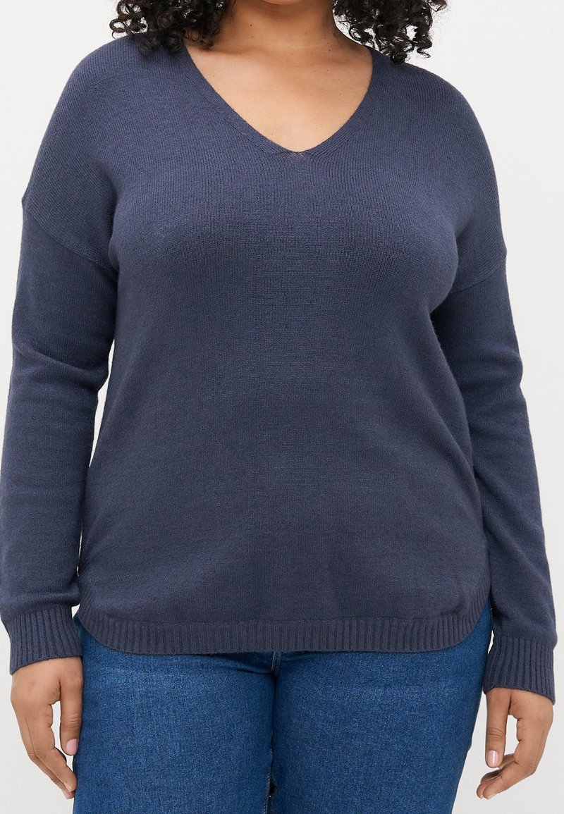 Femme portant un pull à col en V bleu foncé à manches longues et un jean bleu sur un fond clair uni.
