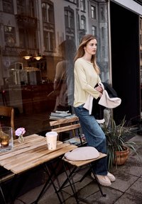 Jeune femme en pull beige et jean tenant une veste, debout près d'une table de café en bois avec un café et une fleur rose.