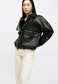 Νεαρή γυναίκα που φοράει μαύρο δερμάτινο μπουφάν με φερμουάρ και μεγάλες μπροστινές τσέπες, καθώς και παντελόνι σε εκρού χρώμα, στέκεται μπροστά σε λευκό φόντο.