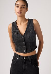 Veste courte en denim noir avec un décolleté en V profond, fermeture à boutons et poches avant ; texture lisse et design ajusté. Associée à une jupe taille haute assortie.