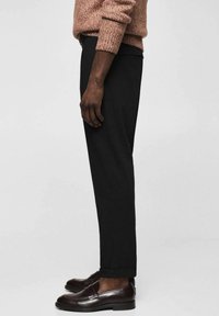 Boční pohled na osobu v černých elegantních kalhotách, hnědých kožených mokasínách a texturovaném hnědém svetr s ohrnutými rukávy.