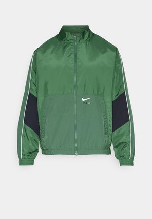 Grøn nylon jakke med lynlås og høj krave, med marineblå detaljer, ribbede manchetter og et lille hvidt logo på forsiden.