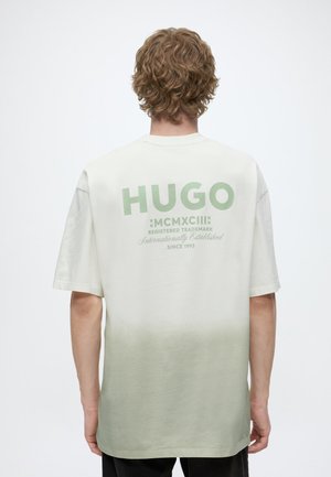 Personne aux cheveux bouclés portant un T-shirt dégradé vert clair et blanc avec "HUGO" et du texte au dos, debout devant un fond uni.