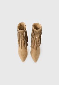 Tan suède enkellaarsjes met puntige neuzen, franjes en verticale rijen zilveren studs op een plain witte achtergrond.
