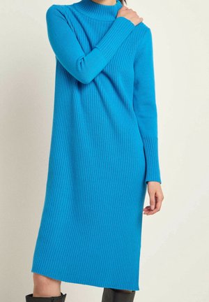 Femme portant une robe-pull midi bleu vif à manches longues côtelées, une main reposant sur l'épaule opposée et des bottes noires.