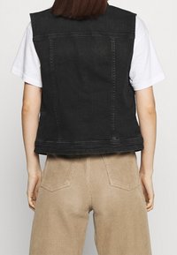 Person med svart ärmlös denimväst över en vit t-shirt och beige kordbyxor, sedd bakifrån mot en enfärgad bakgrund.