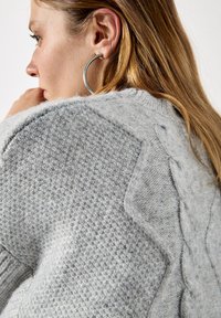 Jasnoszary sweter z dzianiny o teksturowej strukturze, z plecionymi akcentami wzdłuż szwu na ramieniu. Widać dużą srebrną kolczyk w kształcie koła.