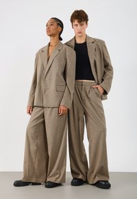 Braon hounds-tooth oversized blejzeri upareni s hlačama širokih nogavica. Glatka tkanina sa strukturiranim ramenima, kontrastne crne majice i robustne crne cipele.