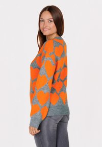 Frau mit langen braunen Haaren, die einen grauen Pullover mit großen orangenen Herzen trägt, kombiniert mit eng anliegenden grauen Jeans, lächelt und schaut über ihre Schulter.