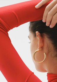 Pendiente de aro dorado con un diseño curvado y un patrón texturizado, llevado en la oreja junto a un pequeño arete de botón, con una blusa de manga larga roja visible.