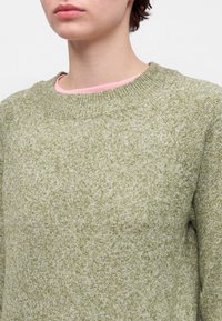Personne portant un pull en maille vert et blanc tacheté avec une chemise rose visible au niveau de l'encolure, montrée du cou au milieu du torse.