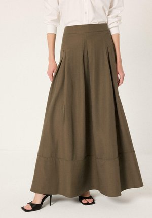 Γυναίκα φοράει μακριά φούστα σε ελιάχρυσο πράσινο με πιέτες, συνδυασμένη με μαύρα πέδιλα με τακούνι και λευκό μακρυμάνικο πουκάμισο.