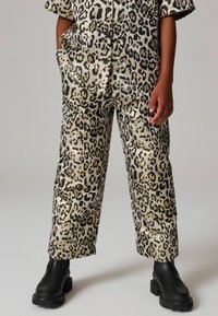 Koszula i spodnie w leopardzi wzór w beżowym i czarnym kolorze, wykonane z luźnego materiału, z krótkimi rękawami i kieszeniami z przodu, zestawione z czarnymi, masywnymi butami.