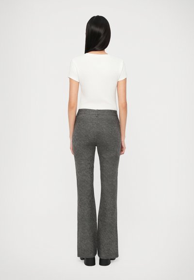 Γυναίκα με μακριά ίσια μαλλιά, που φοράει ένα λευκό μπλουζάκι με κοντά μανίκια και γκρι υφασμάτινο παντελόνι με ψηλή μέση και φούντα, στέκεται με την πλάτη στραμμένη προς τα εμπρός.