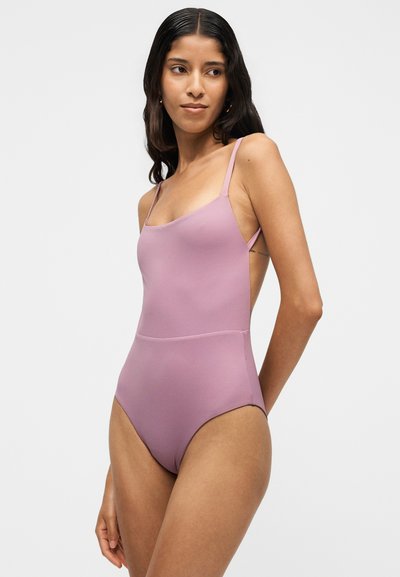 Femme aux longs cheveux foncés portant un maillot de bain une pièce sans manches de couleur violet clair, debout devant un fond clair uni.