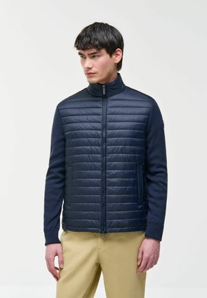 Man wearing navy quilted zip-up jacket with knitted sleeves and beige pants, standing against plain light background, looking left.