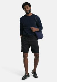 Marineblaues Langarmhemd, schwarze Shorts und schwarze Slipper. Model hält eine dunkle Tasche und steht vor einem schlichten weißen Hintergrund.