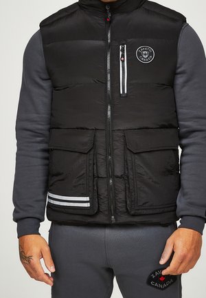 Black quilted vest with front zipper, chest pocket, and two flap pockets worn over dark gray long-sleeve top and matching pants.