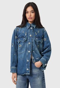 Keskmise sinise tooniga teksajakk, millel on lillemustrilised kaunistused ja eest nööbitav sulgus. Varustatud kahe rinnataskuga ja lahtise istuvusega.