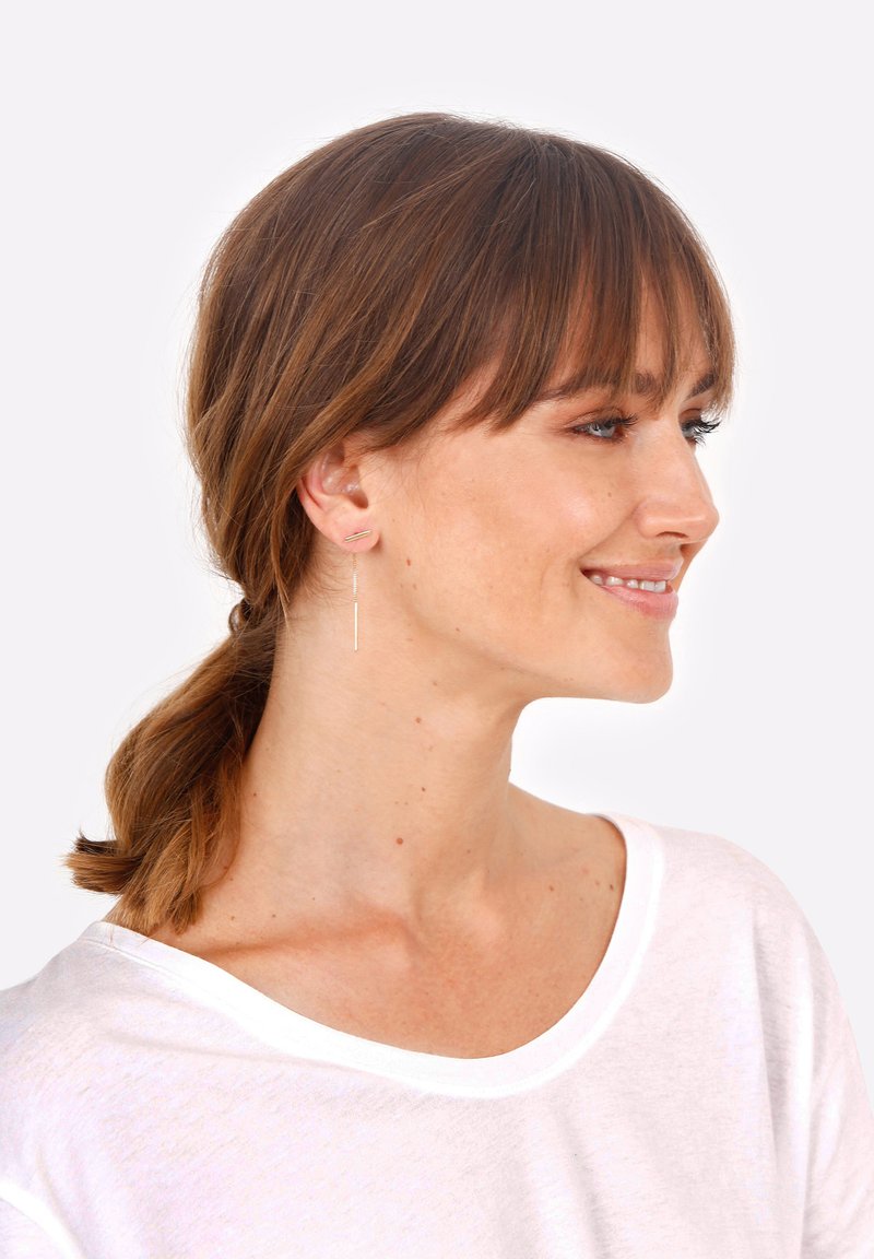 Pendientes de plata con un diseño de varilla delgada, que presentan un pequeño detalle de barra horizontal, llevados por una persona con cabello castaño de longitud media.