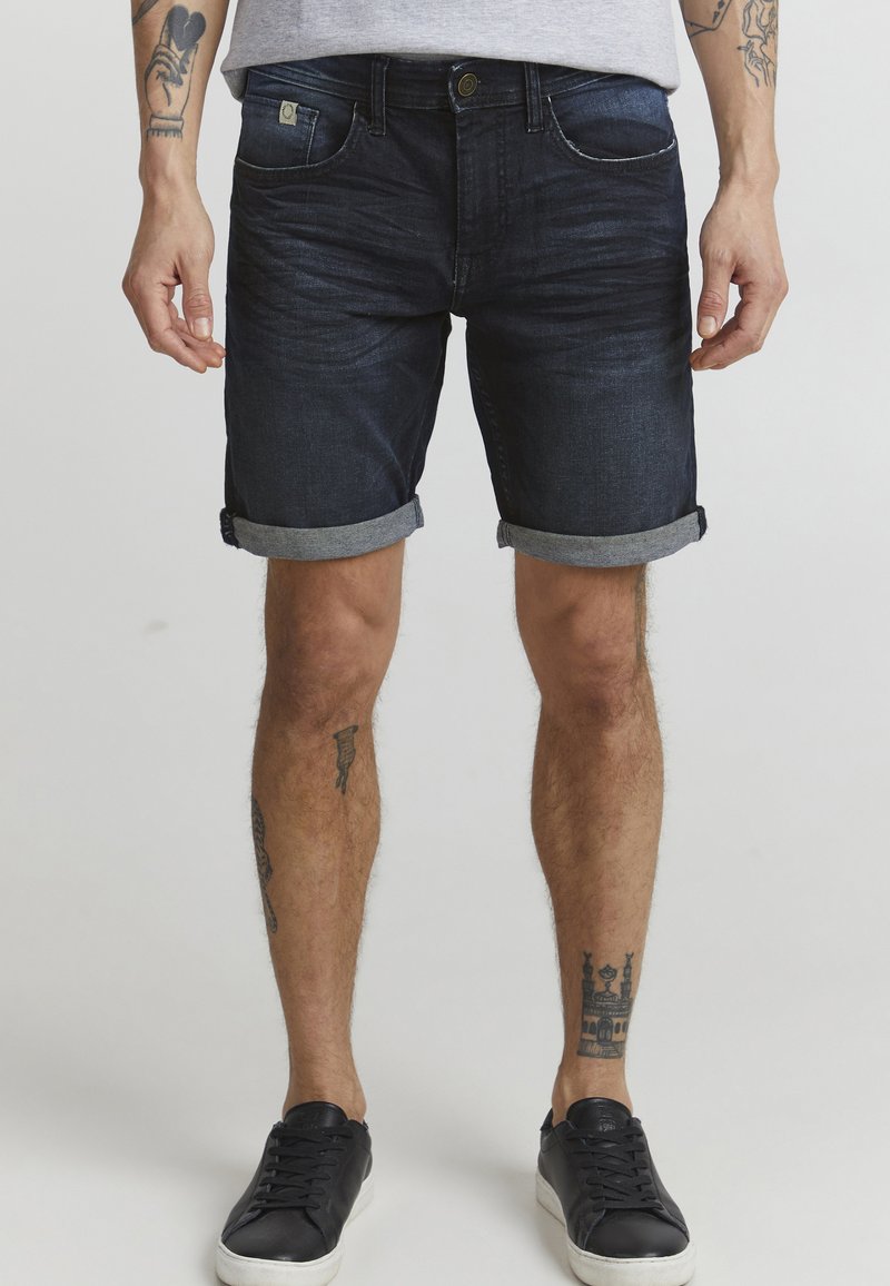 Temno modri denim kratki hlače s zvitim robom, ki vključujejo rahlo zbledelost in sprednje žepe. V kombinaciji z črnimi teniskami in sivo majico.