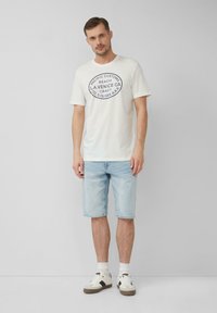Witte katoenen T-shirt met een ronde marineblauwe grafische print, gecombineerd met lichte denimshorts en witte sneakers met zwarte accenten.