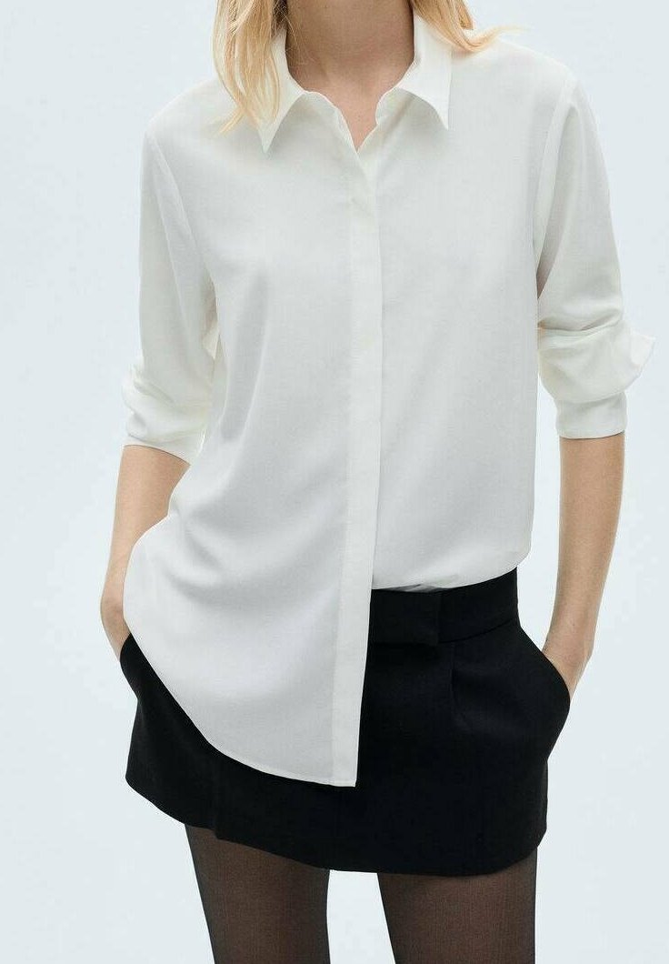 Femme portant une chemise blanche boutonnée avec manches retroussées, jupe noire et collants noirs transparents, mains dans les poches de la jupe, sur fond uni.