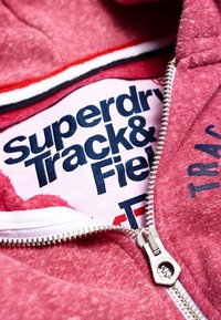 Giacca rossa con zip parzialmente aperta, che rivela una camicia bianca con il testo in grassetto blu navy "Superdry Track & Field" all'interno.
