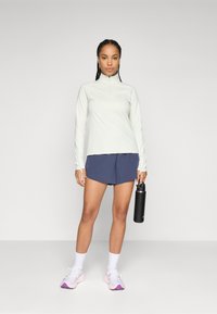 Light gray long-sleeve top with a zip collar, paired with navy shorts. Black water bottle held in right hand; pink and white sneakers.
