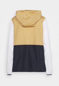 Färgblockerad jacka med en beige överdel, vita ärmar och en marinblå nederdel. Tillverkad av lätt, vattentåligt material med huva.