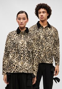 Leopárdmintás kabátok fekete szegéllyel, gombos záródással és elülső zsebekkel rendelkeznek. Két személy viseli őket egy egyszínű háttér előtt.