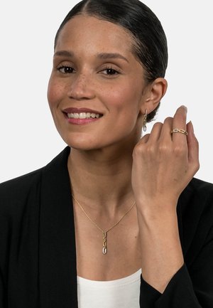 Gold chain necklace with a pendant, gold hoop earrings, and a gold ring with a small accent. Model wears a black blazer over a white top.